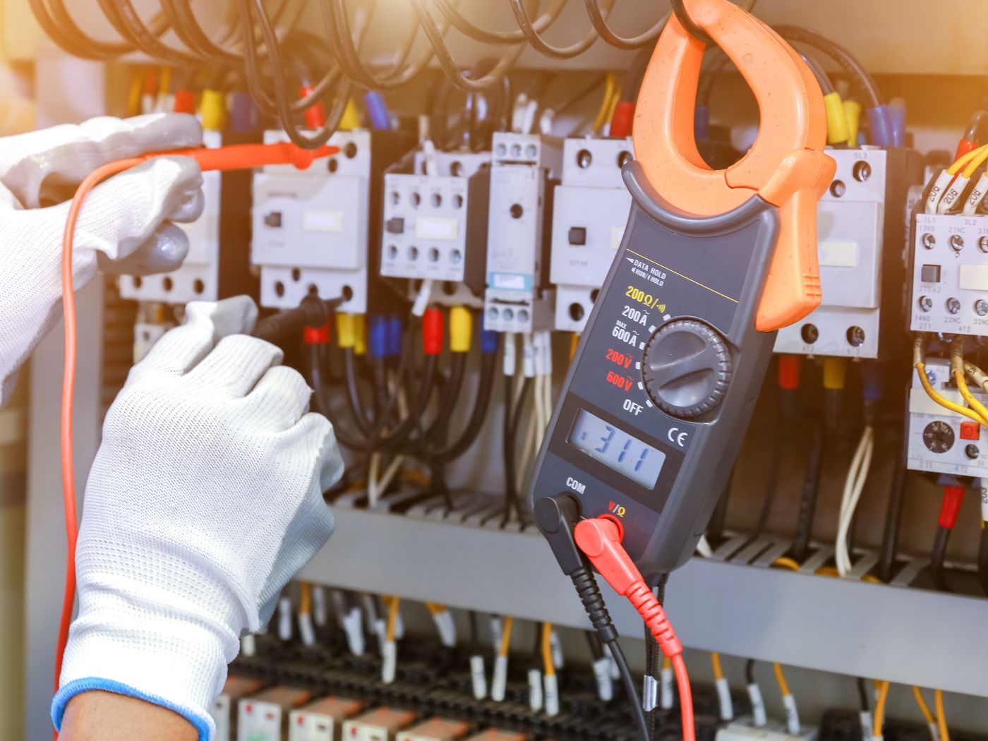Gloved hands using a clamp meter to test a switchboard full of contactors and breakers