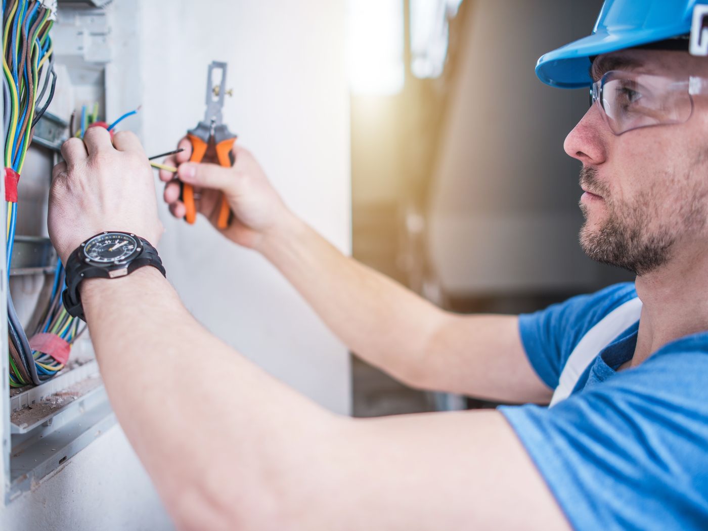 An electrician in safety glasses and a blue shirt wiring a wall box with a screwdriver
