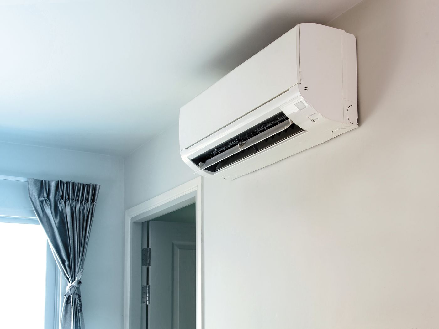A white high-wall heat pump above a doorway in a bright room with a curtained window