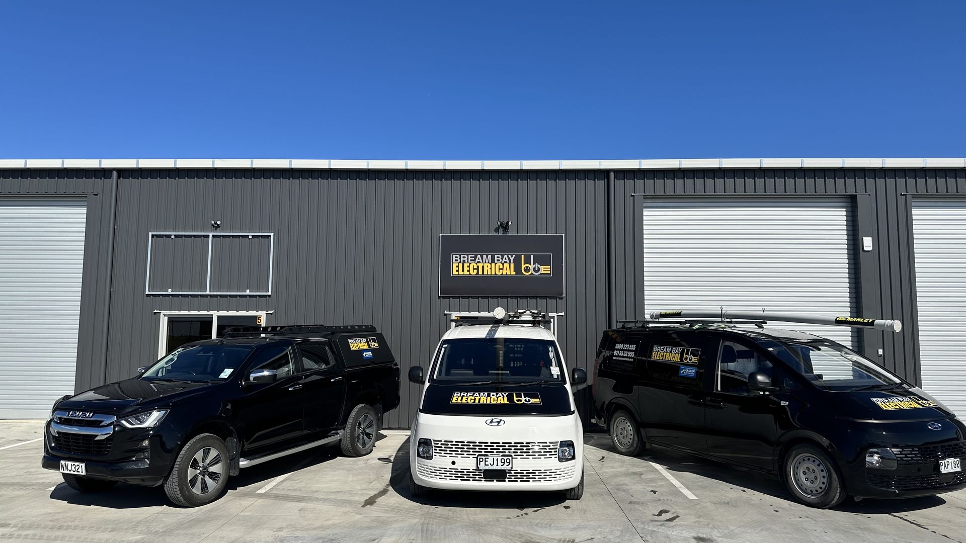 Three Bream Bay Electrical vehicles, a black ute, a white van and a black van, parked outside the company's grey workshop unit with its sign above the door