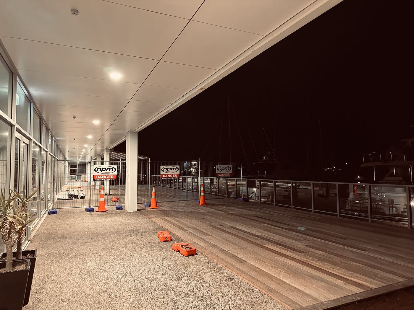 A covered marina walkway at night lit by rows of ceiling downlights, with a timber boardwalk and safety cones where work is under way
