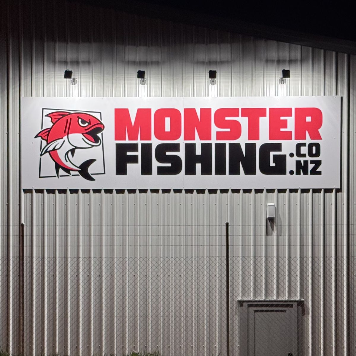 The Monster Fishing sign on a steel-clad building at night, lit from above by a row of floodlights
