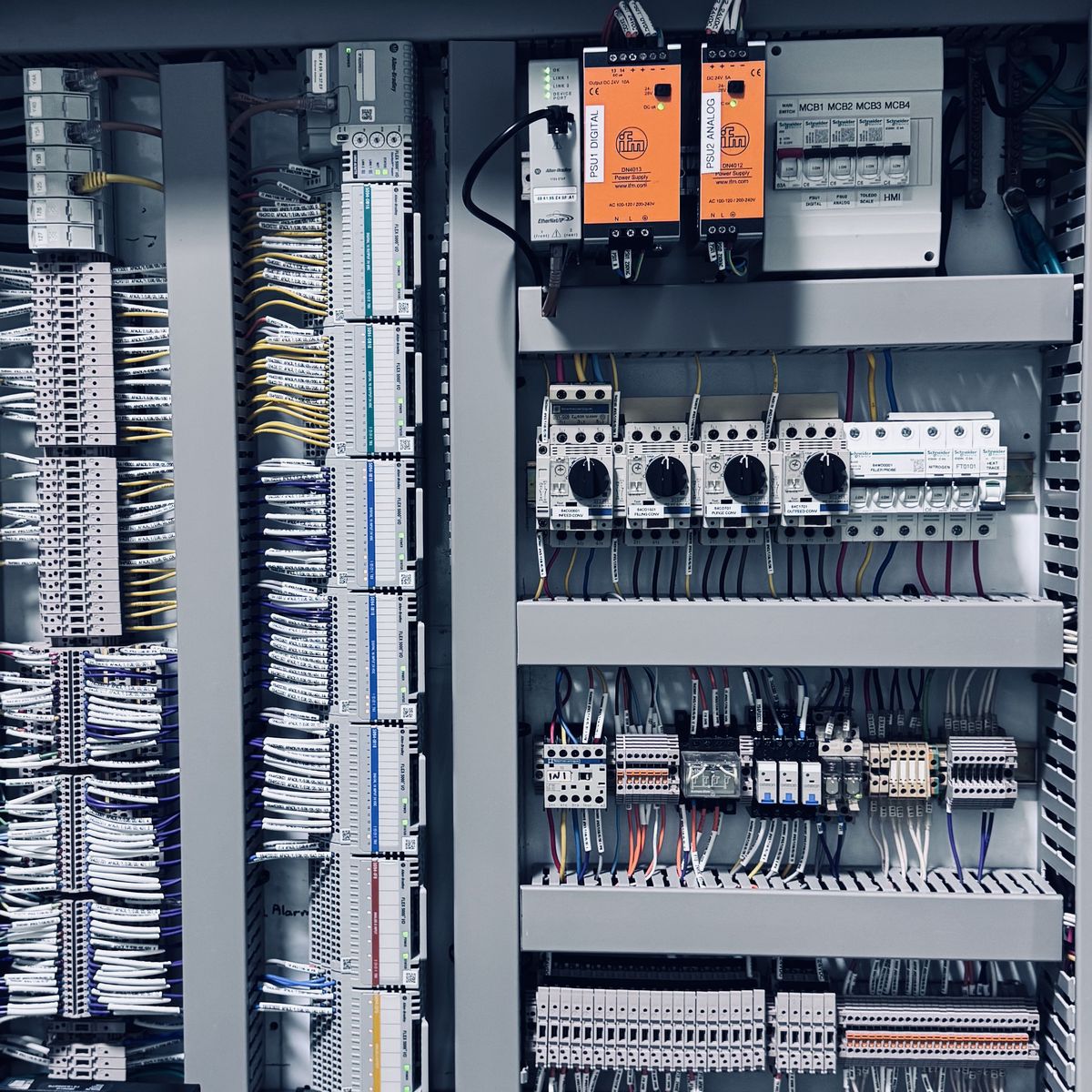 Inside an industrial control cabinet: rows of terminals with labelled wiring on the left, two orange power supplies, motor protection breakers and contactors on DIN rail