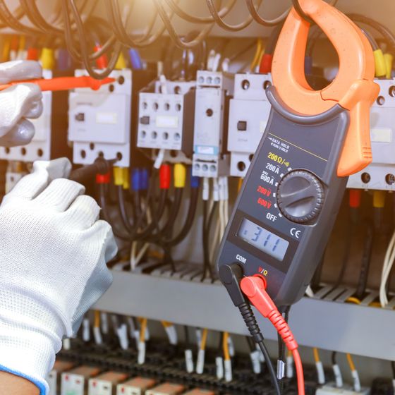 A white-gloved hand holding an orange clamp meter against the contactors in a switchboard