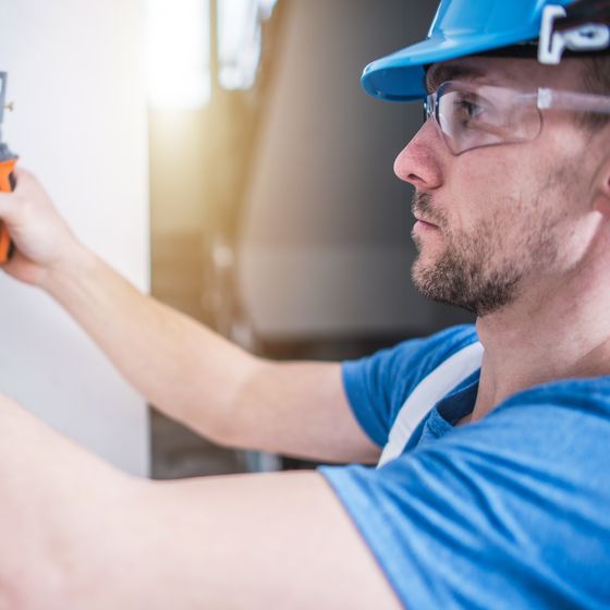 An electrician in a blue hard hat and safety glasses reaching up to work at a wall