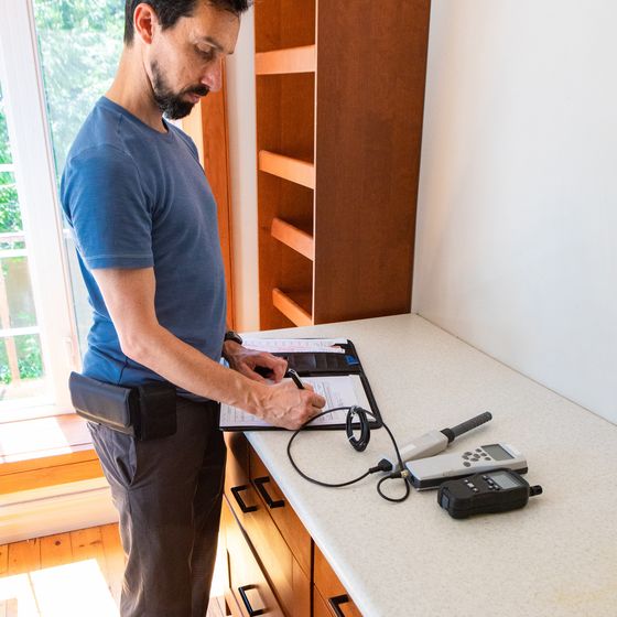 A man in a blue t-shirt writing on a clipboard at a bench, with two handheld test instruments beside him