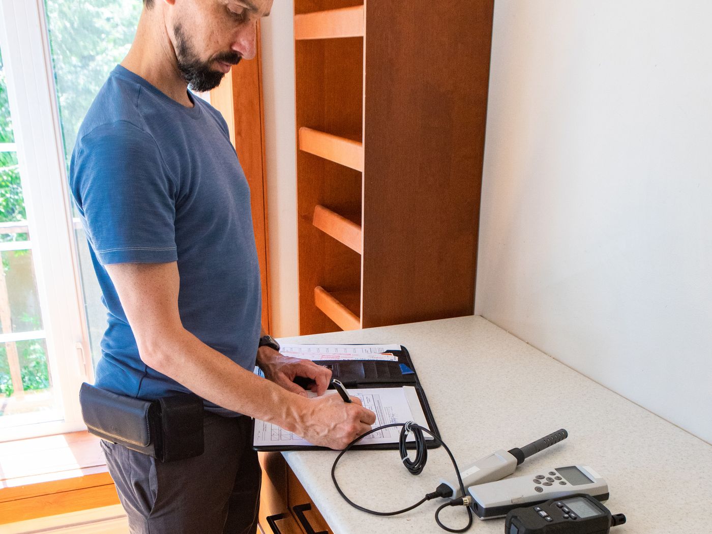 A man writing up results on a clipboard at a bench, with handheld test instruments and a coiled lead next to the paperwork